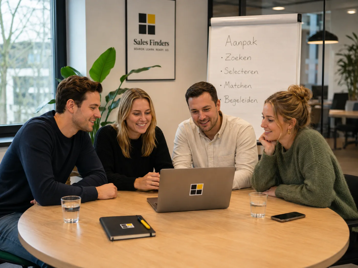 Vier collega’s van Sales Finders kijken samen naar een laptop aan een ronde tafel. Op de flip-over ernaast staat de aanpak: zoeken, selecteren, matchen en begeleiden.