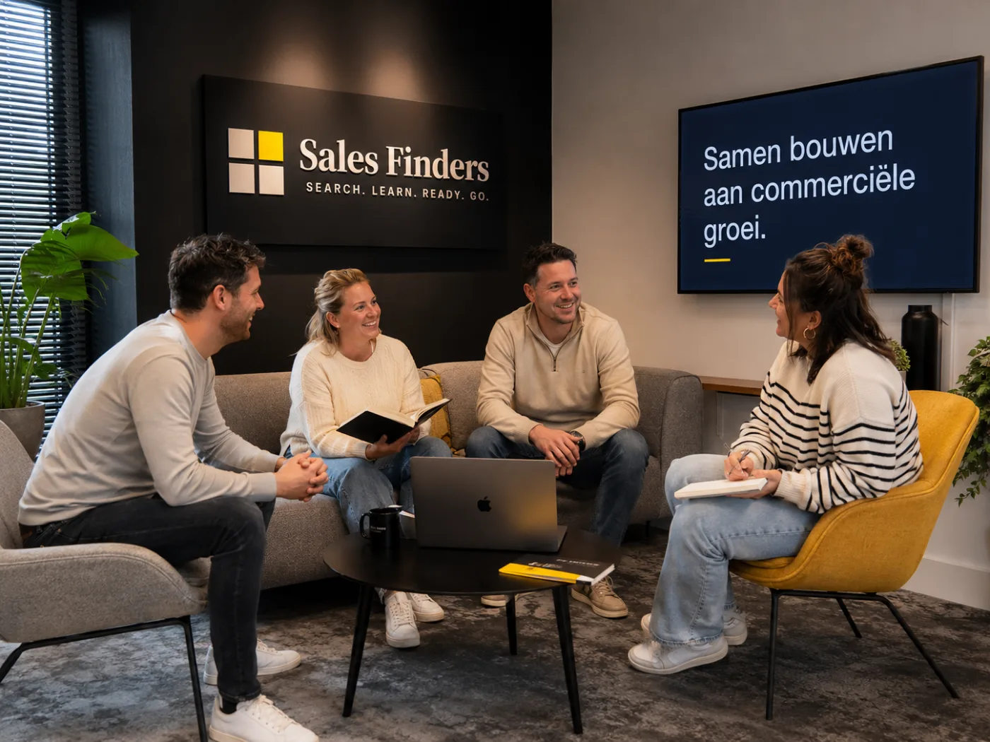 Vier collega’s van Sales Finders in gesprek in de zithoek bij het logo, met op het scherm achter hen de zin samen bouwen aan commerciele groei.