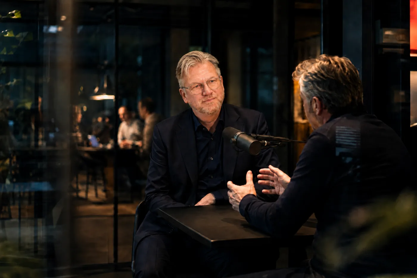 Twee mannen nemen een aflevering van Salesbazen op aan een statafel met een podcastmicrofoon ertussen. De een luistert aandachtig, de ander praat met zijn handen.